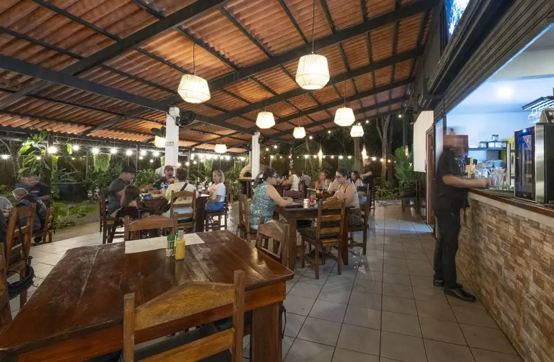 Salón al aire libre con techo de madera, lámparas colgantes, mesas de madera y comensales