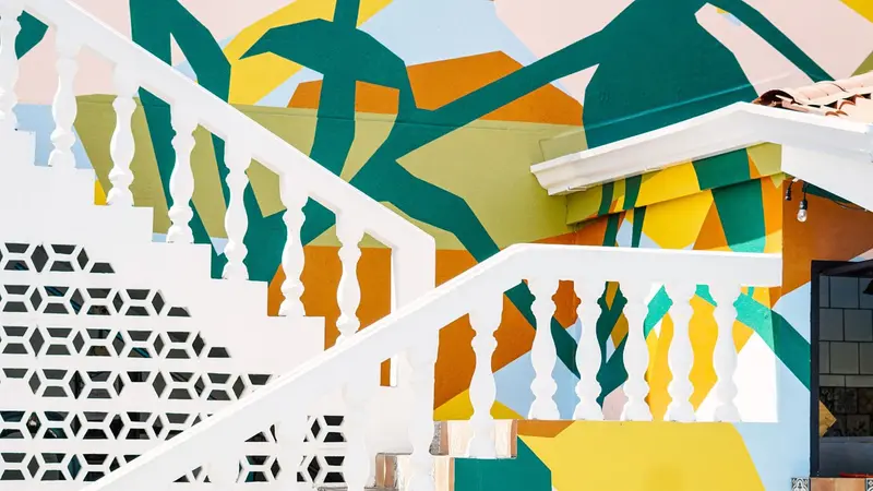 Escalera blanca con barandas torneadas frente a un mural geométrico de hojas amarillas, verdes y naranjas