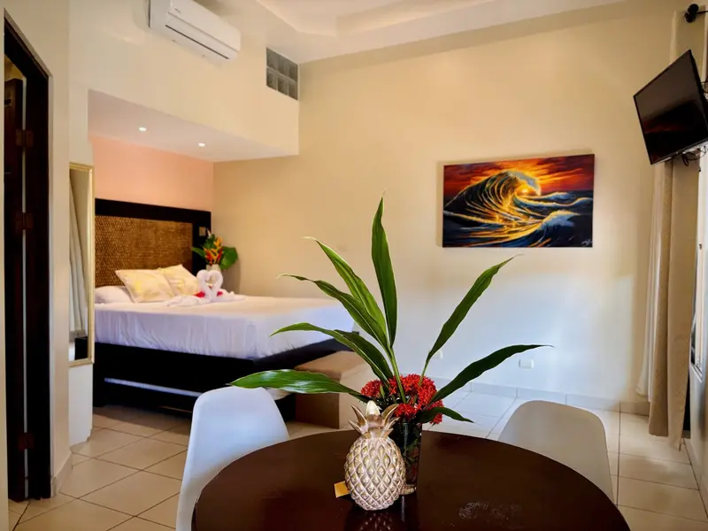 Suite con cama y toallas dobladas en forma de cisnes, mesa con sillas blancas y una piña decorativa, y una pintura de una ola al atardecer