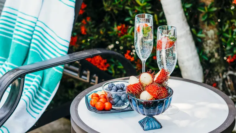 Dos copas de espumante y un cuenco de fruta con fresas y rambután sobre una mesa junto a la piscina