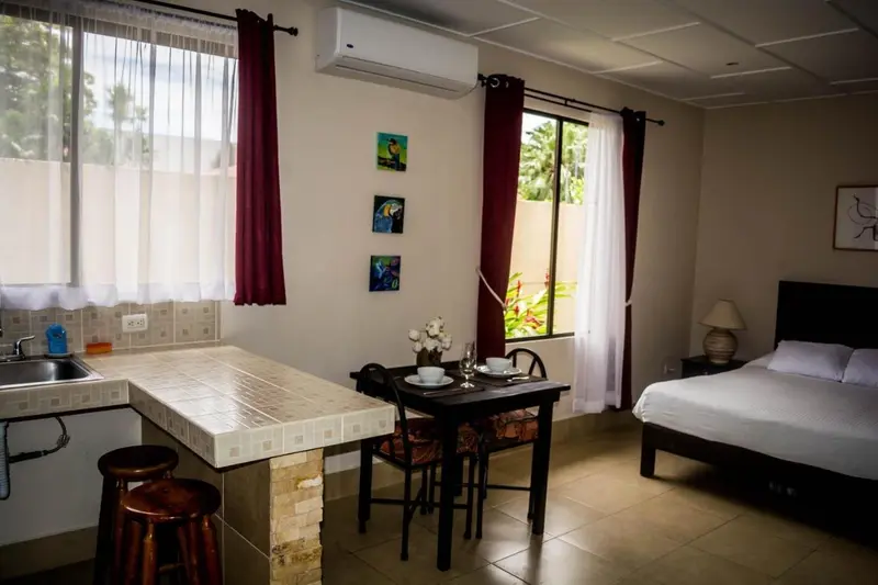Habitación con cama, cocina pequeña con barra y mesa de comedor frente a ventanas