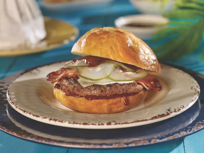 Hamburguesa en pan brioche con tocineta y pepinillos, servida en un plato blanco con borde rústico