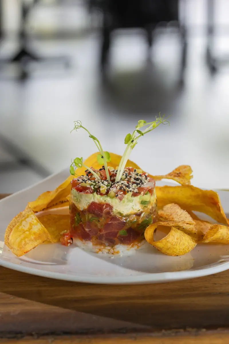Torre de atún con arroz de sushi y guacamole sobre un plato blanco, rodeada de chips de plátano