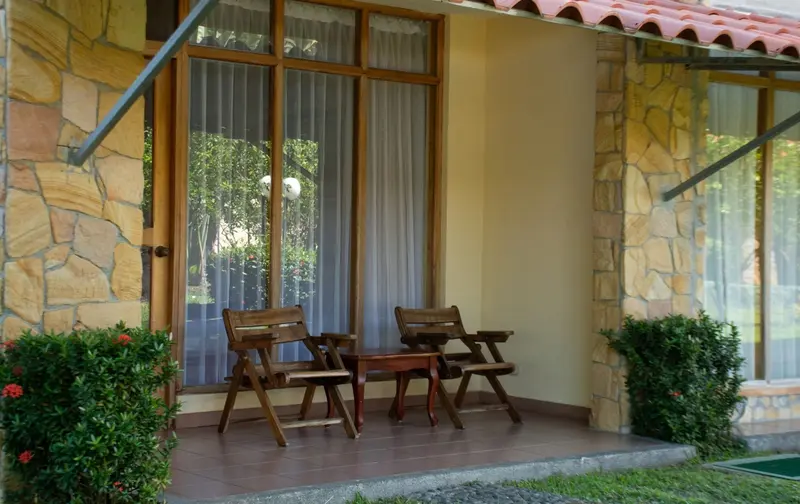 Terraza de una habitación con dos sillas de madera y una mesa pequeña, entre columnas de piedra y plantas
