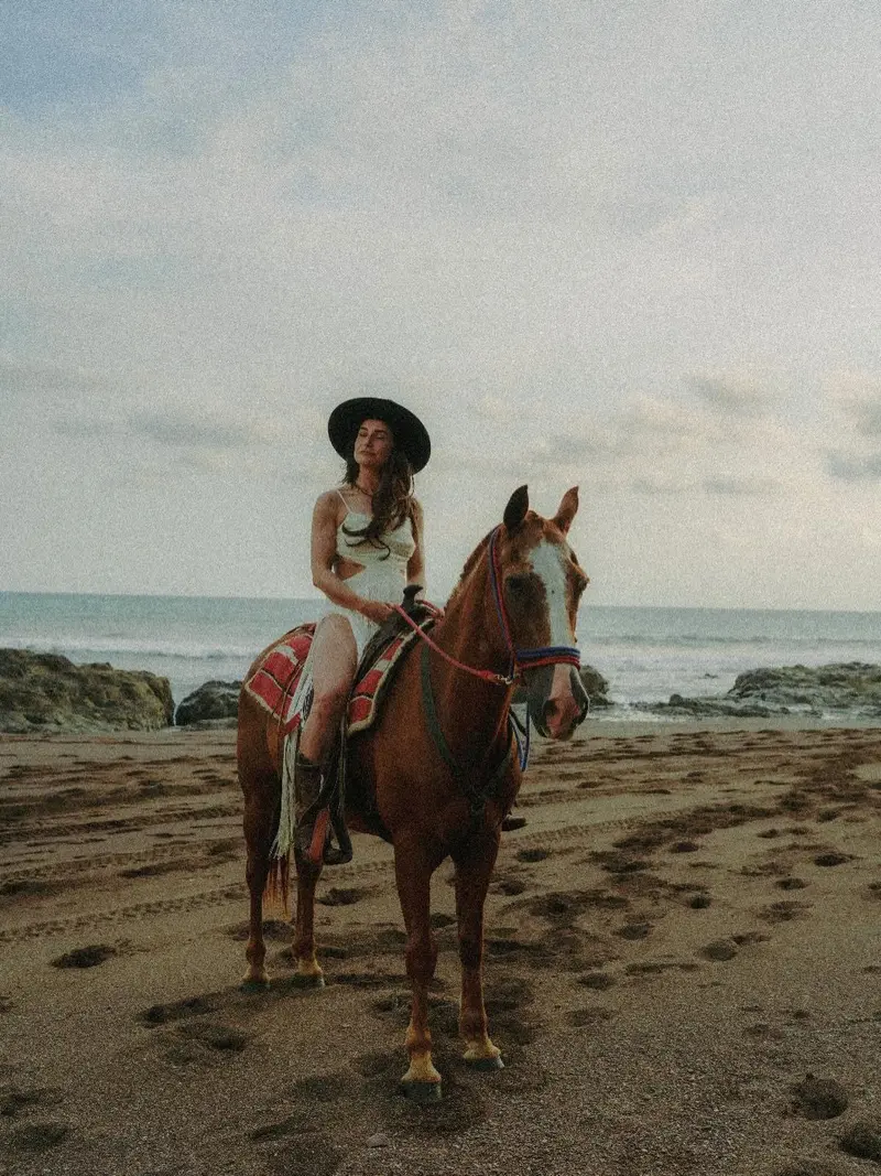 Jinete con sombrero sobre un caballo castaño en una playa de arena oscura, con rocas y mar al fondo