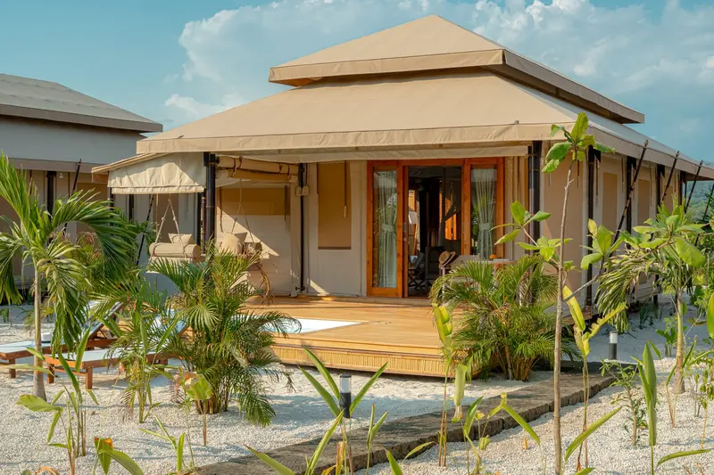 Villa tipo tienda de campaña con techo de paja piramidal y terraza de madera, entre plantas de playa