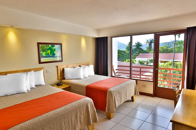 Habitación con dos camas dobles, cobertores naranja y puertas de vidrio hacia un balcón