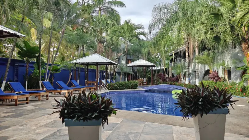 Piscina del hotel con camastros azules, cabanas y el edificio de habitaciones al fondo