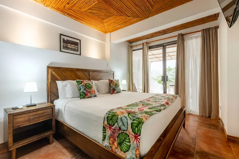 Habitación con cama king de cabecera de madera, cielo raso de bambú y puertas de vidrio hacia el balcón
