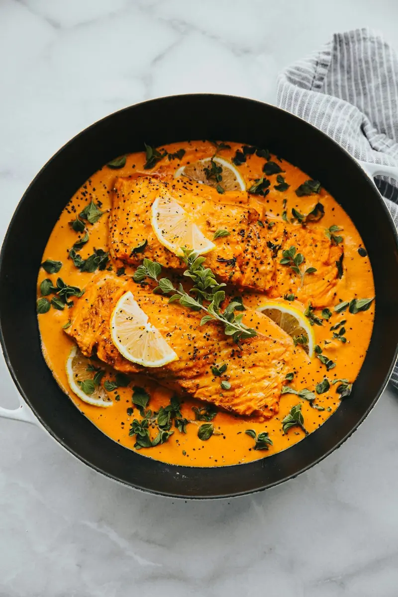 Salmón en salsa de curry naranja con rodajas de limón y hierbas, en una sartén negra sobre mármol
