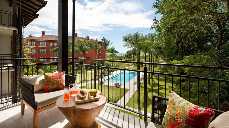 Balcón con sofá, mesa baja y barandas de metal con vista a la piscina y los jardines