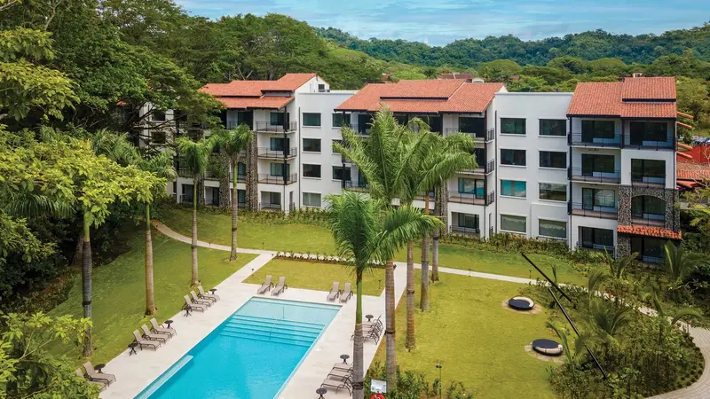Edificio de villas de cuatro pisos con techos de teja entre palmeras y jardines, con la piscina en primer plano