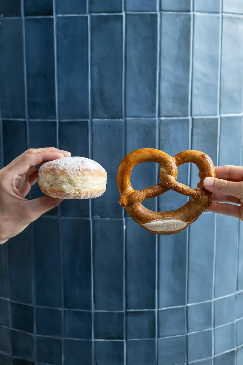 Pretzel alemán y un pan redondo sostenidos frente a una pared de azulejos azules