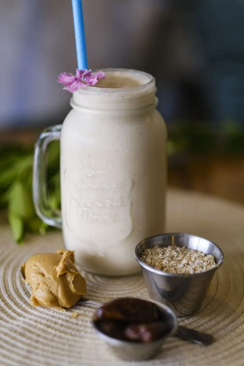 Batido en un frasco con pajilla azul y una flor, junto a mantequilla de maní, avena y dátiles