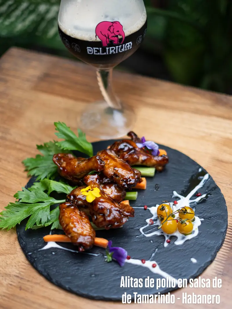 Alitas de pollo glaseadas con flores comestibles sobre una pizarra, con una copa de cerveza al fondo