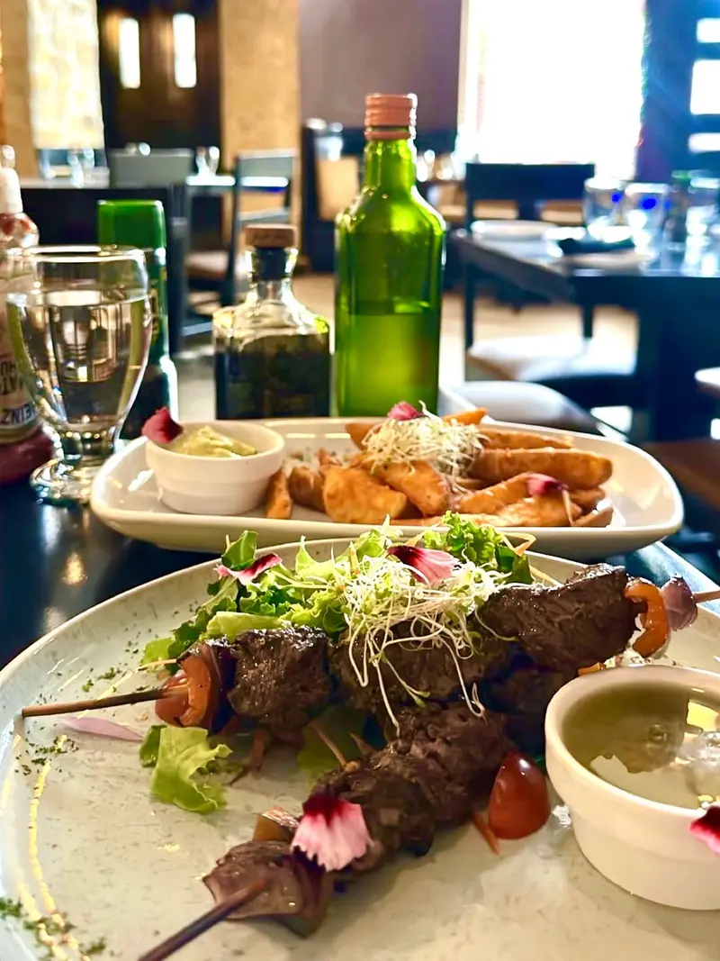 Pinchos de carne a la parrilla sobre hojas verdes y germinados, con salsa verde, y una fuente de papas al fondo