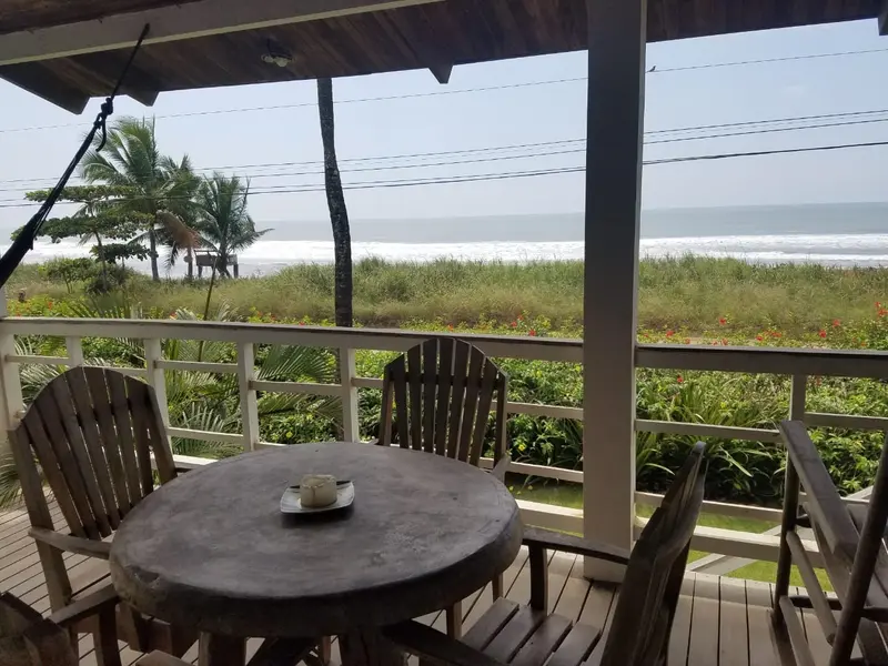 Balcón techado con mesa redonda y sillas frente al mar, con palmeras y oleaje al fondo