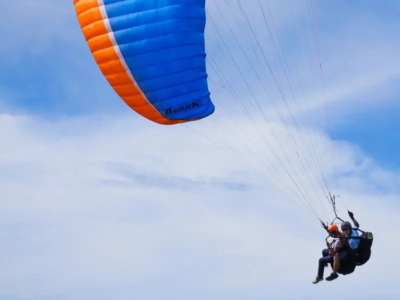 Ala de parapente azul y naranja vista desde abajo contra el cielo, con un tándem en el arnés