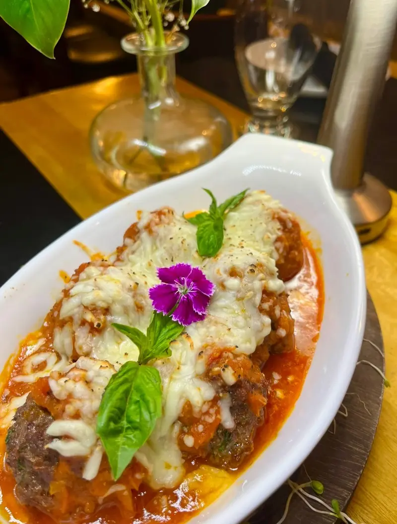 Albóndigas con salsa de tomate y queso gratinado en una fuente blanca, con albahaca y una flor morada