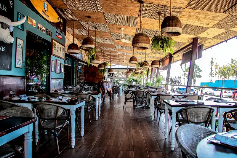 Terraza techada con cielo raso de bambú, mesas servidas, lámparas de mimbre y la playa al fondo