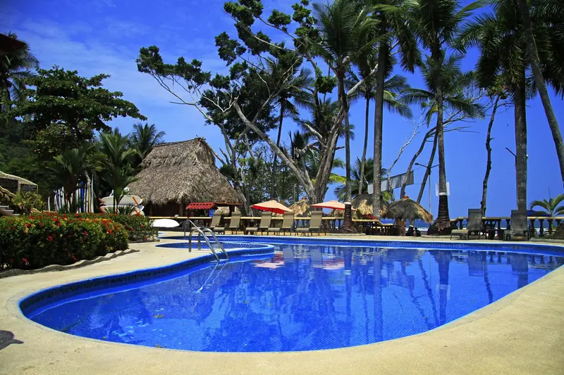 Piscina de forma curva rodeada de palmeras, con sombrillas y un rancho de paja al fondo