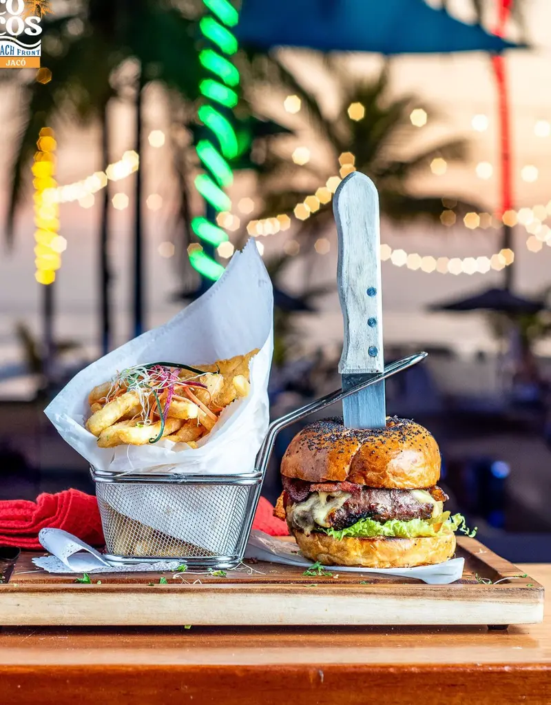 Hamburguesa con papas en cono de papel sobre una tabla de madera, con luces y el mar al atardecer