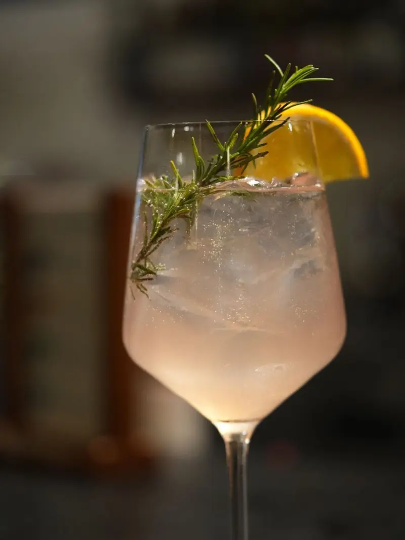 Copa de cóctel rosado claro con una rodaja de naranja y una rama de romero