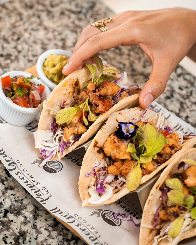 Tacos con relleno frito y repollo morado sobre papel con el logo del restaurante, con pico de gallo y guacamole al lado