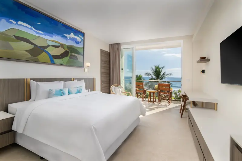 Habitación con cama king, un cuadro sobre la cabecera y un ventanal con vista al mar y a las palmeras