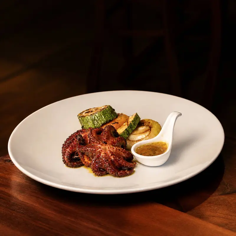Pulpo a la parrilla con rodajas de zucchini y una cucharita con salsa amarilla