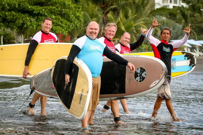 Instructores y alumnos con licras saliendo del agua con sus tablas, sonriendo y levantando los brazos
