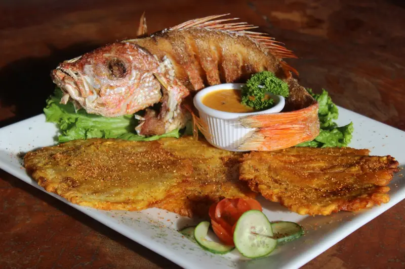 Pargo entero frito con patacones, ensalada y salsa caribeña en un plato blanco