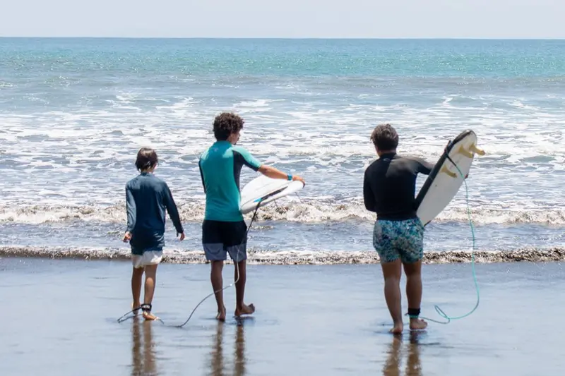 Tres personas de espaldas caminando hacia el mar con tablas, sobre la arena mojada