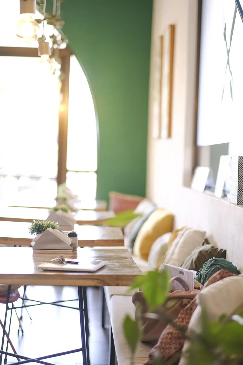 Salón con mesas de madera, un banco con almohadones y una pared verde junto a una ventana en arco