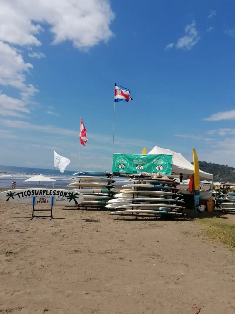 Puesto de la escuela en la playa con carpa, tablas en el suelo y banderas bajo un cielo azul