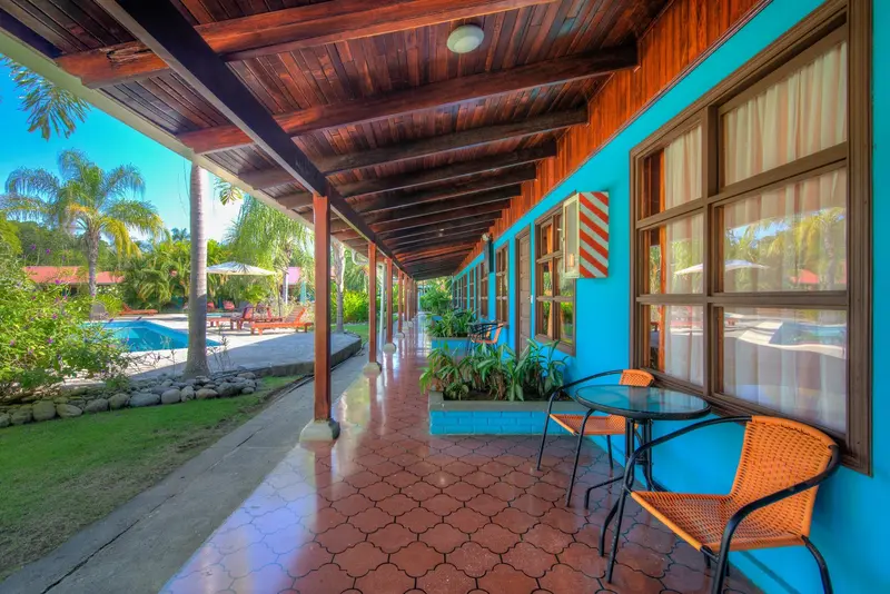 Corredor con mesa y sillas frente a las habitaciones azules, con piscina y palmeras al fondo