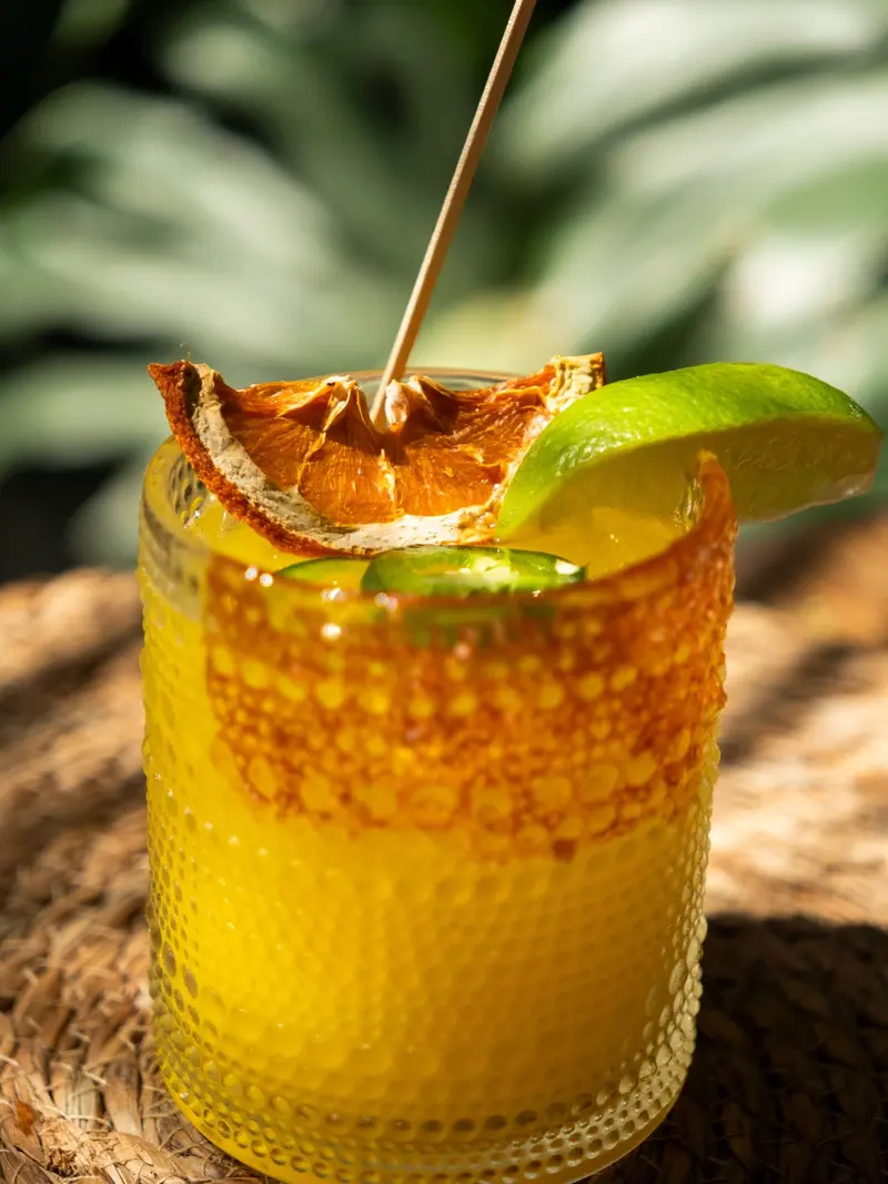 Cóctel amarillo con una rodaja de naranja deshidratada y una lima