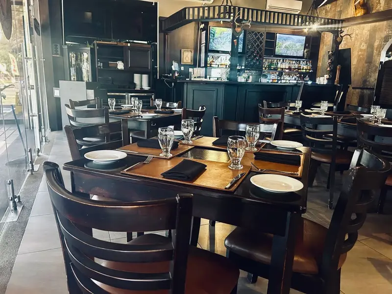 Salón con mesas de madera oscura, sillas de respaldo de listones y la barra con botellas al fondo