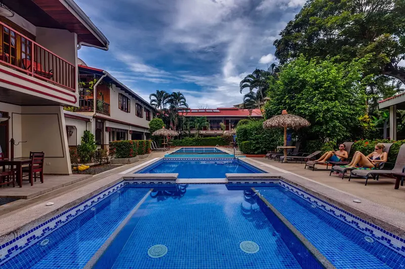 Patio con dos piscinas azules, techos de teja roja, sombrillas de palma y árboles alrededor