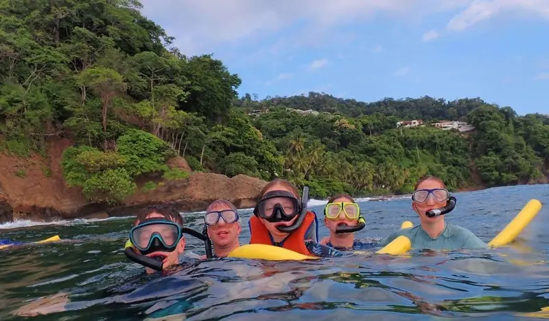 Grupo con máscaras de snorkel en el agua frente a una costa con selva, con fideos de espuma amarillos