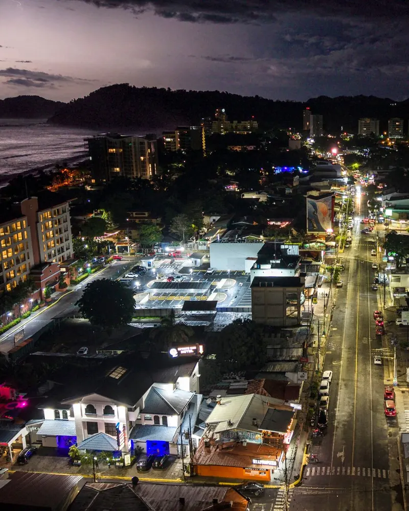 Vista aérea de Jacó al anochecer, con la avenida Pastor Díaz iluminada, edificios junto a la playa y el océano Pacífico.