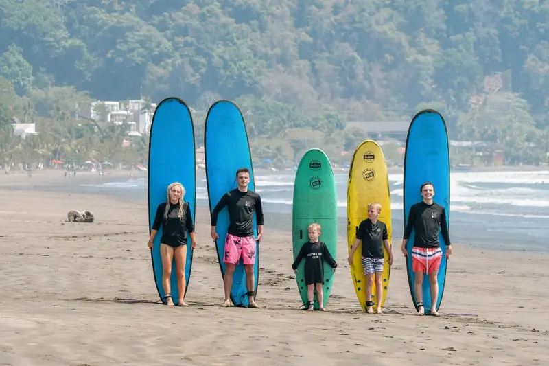 Cinco personas, dos de ellas niños, de pie en la arena junto a tablas de surf azules, una verde y una amarilla