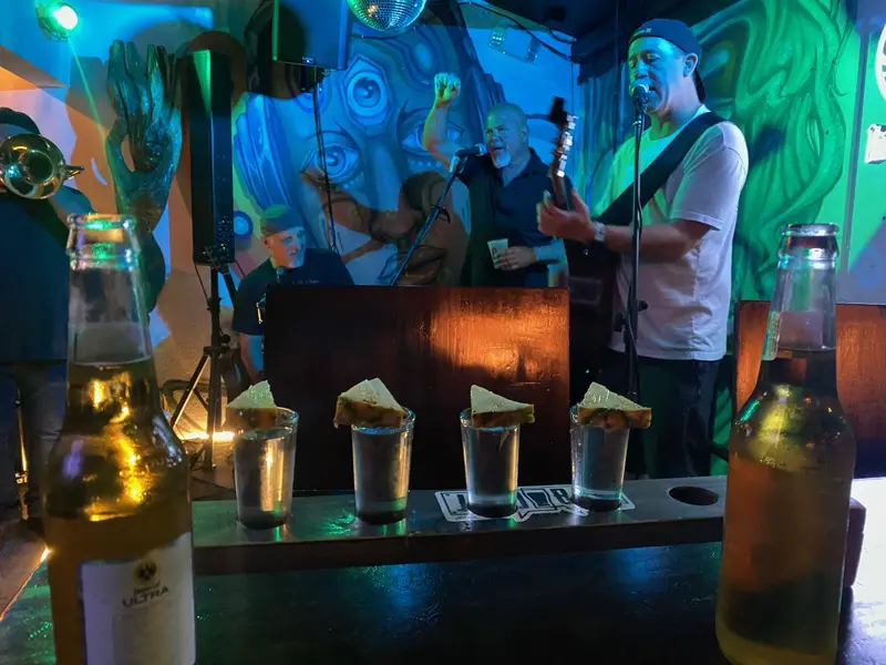 Banda en el escenario detrás de una tabla con cuatro shots con piña y dos botellas de cerveza