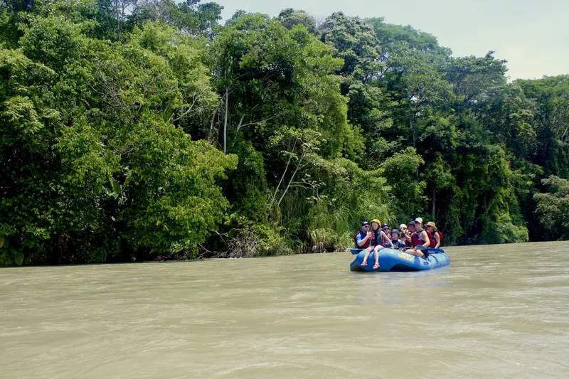 Balsa azul con rafters a lo lejos sobre un río ancho entre selva verde