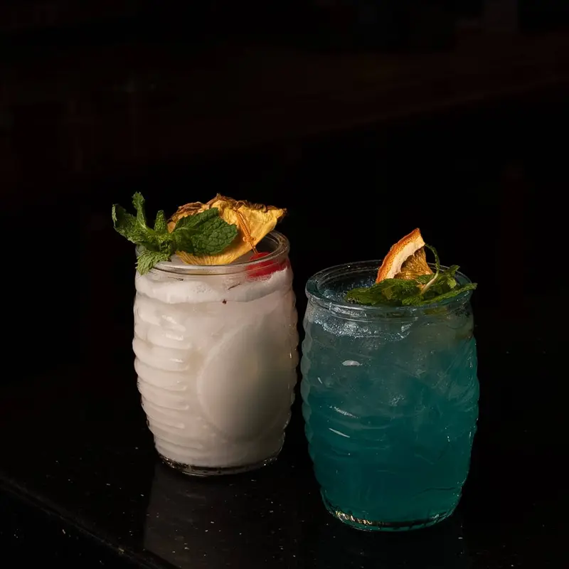 Dos cócteles en vasos con relieve, uno blanco y otro azul, con menta y naranja deshidratada