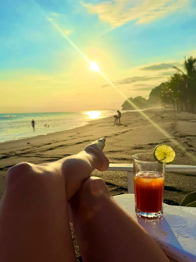 Sunset over the beach with the sun low, a glass with a lime slice in the foreground and people walking along the shore