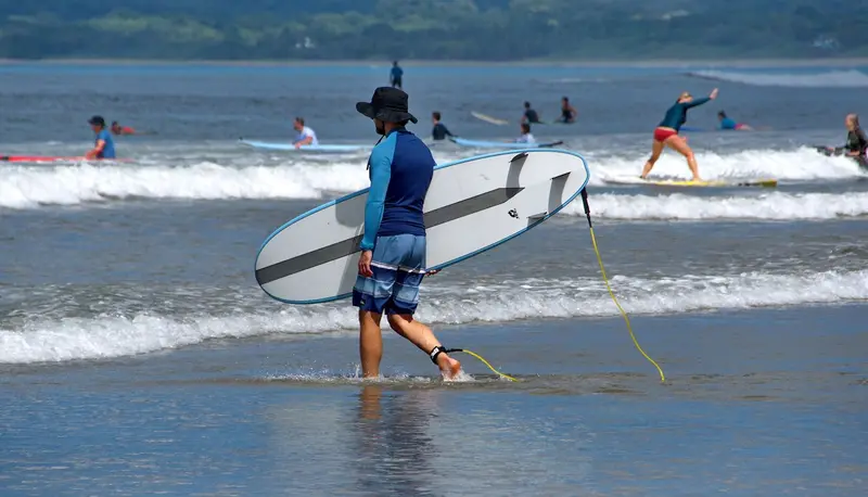 Surfista con camiseta de protección sale del mar cargando una tabla de surf con la correa, con más surfistas al fondo