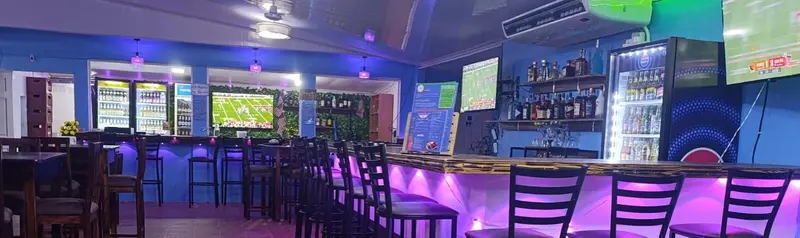 Barra iluminada en morado con taburetes, neveras de bebidas y pantallas con un partido