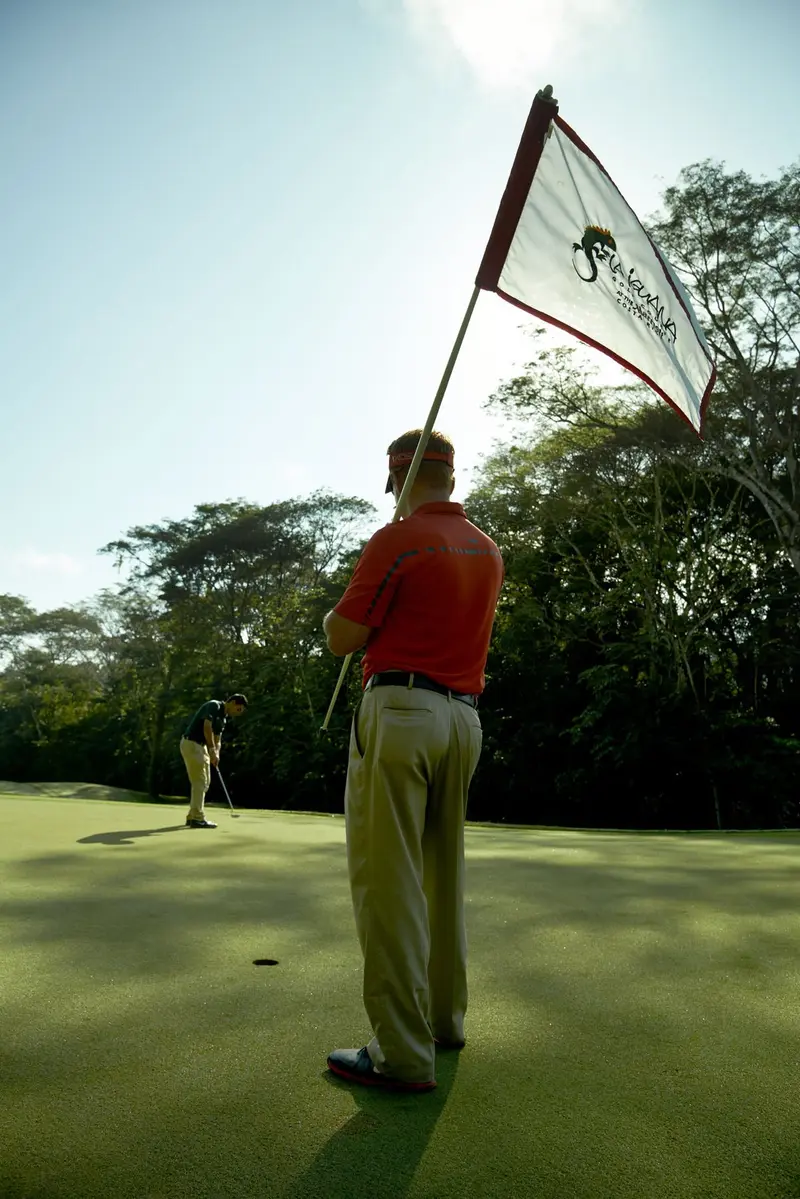 Golfista de camisa roja sosteniendo la bandera blanca de un hoyo, a contraluz