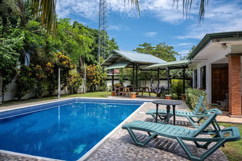 Piscina azul con camastros y un rancho de techo de lámina entre palmeras y jardín